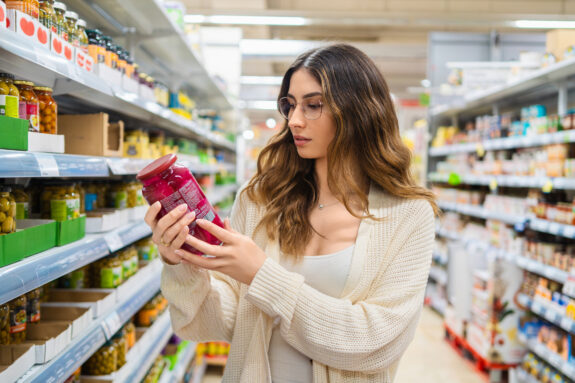Young adult woman with glasses examining a jar in a grocery store aisle